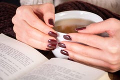 Woman Hands With Cup Of Tea And Brown Manicure On Finger Nails And Open Book On Wooden Retro Desk Stock Photos