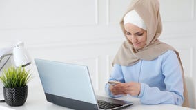Woman in hijab is reading something on mobile phone sitting on her working place with laptop. stock footage Woman in hijab is reading something on mobile phone sitting on her working place with laptop. stock footage
