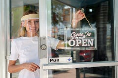 Smiling woman in white shirt wearing face shield hanging open sign for storefront business on the door