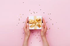 Womans hands holding gift or present box decorated confetti on pink pastel table top view. Flat lay composition for birthday.