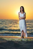 Wonderful girl wearing white long dress standing on sea sunset sideview. Deep blue sea waves behind her. Breezing fresh sea breeze. Watching on sun going down Stock Image Wonderful girl wearing white long dress standing on sea sunset sideview. Deep blue sea waves behind her. Breezing fresh sea breeze. Watching on sun going down Stock Image