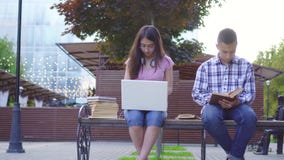 Young asian students man with a book and a woman with a laptop sitting on a bench stock footage Young asian students man with a book and a woman with a laptop sitting on a bench stock footage