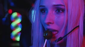 Young beautiful blonde woman gamer talking in the mic of her headphones and licking a lollipop
