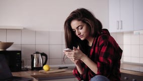 Young beautiful brunette girl with messy hair in flannel shirt sitting in her kitchen and texting someone. stock footage Young beautiful brunette girl with messy hair in flannel shirt sitting in her kitchen and texting someone. stock footage