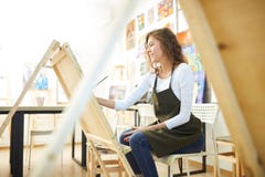 Young beautiful girl with curly hair dressed in white blouse, brown apron and jeans paints a picture at the easel in the royalty free stock photo Young beautiful girl with curly hair dressed in white blouse, brown apron and jeans paints a picture at the easel in the royalty free stock photo