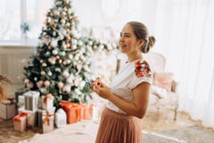 Young beautiful mother holds New Year`s decorations standing in the full of light cozy room next to the New Year`s tree stock photo Young beautiful mother holds New Year`s decorations standing in the full of light cozy room next to the New Year`s tree stock photo