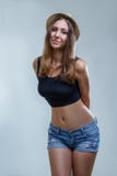 Young Beautiful Shy Woman In Hat, Denim Shorts And Black Shirt On Gray Background In Studio Royalty Free Stock Photos