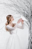 A young blonde bride in white wedding dress on a background of white walls and white tree in the background holds a Stock Photo A young blonde bride in white wedding dress on a background of white walls and white tree in the background holds a Stock Photo