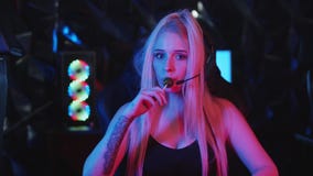 Young blonde woman gamer in big headphones licking a lollipop and looking in the camera