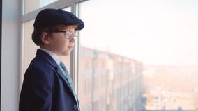 Young boy in business suit, tie and cap looks out from window in business office stock video footage Young boy in business suit, tie and cap looks out from window in business office stock video footage