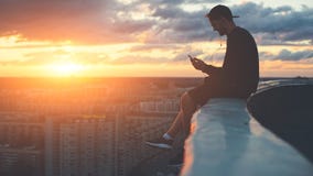 Young brave man sitting on the edge of the roof with smartphone at sunset royalty free stock photography Young brave man sitting on the edge of the roof with smartphone at sunset royalty free stock photography