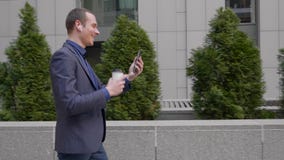 A young businessman walking down the street with wireless earphones in the ears and happily communicates on the video call stock footage A young businessman walking down the street with wireless earphones in the ears and happily communicates on the video call stock footage