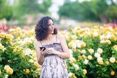 Young Caucasian woman with dark curly hair near yellow rose bush in a rose garden with black smartphone, reading emails, checking stock photography Young Caucasian woman with dark curly hair near yellow rose bush in a rose garden with black smartphone, reading emails, checking stock photography