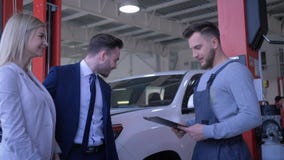 Young couple clients consults with worker of auto repair shop about car maintenance and write assignments on tablet at stock video footage Young couple clients consults with worker of auto repair shop about car maintenance and write assignments on tablet at stock video footage