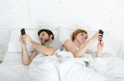 Young couple using mobile phone in bed ignoring each other in relationship communication problems. Sad view of young married couple using their mobile phone in Young couple using mobile phone in bed ignoring each other in relationship communication problems. Sad view of young married couple using their mobile phone in