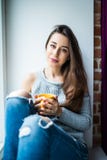 Young girl enjoying an ideal day sitting in the window-sill. Wearing casual denim jeans and blouse, crossing legs. And looking through the window Stock Photo Young girl enjoying an ideal day sitting in the window-sill. Wearing casual denim jeans and blouse, crossing legs. And looking through the window Stock Photo