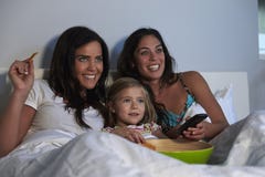 Young girl watching TV in bed with gay female parents Stock Photos Young girl watching TV in bed with gay female parents Stock Photos