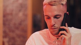 Young handsome man talking on the smartphone in the night cafe. He sitting near neon signage and drinking cocktail stock video Young handsome man talking on the smartphone in the night cafe. He sitting near neon signage and drinking cocktail stock video