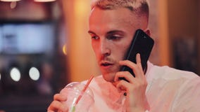 Young handsome man talking on the smartphone in the night cafe. He sitting near neon signage and drinking cocktail stock footage Young handsome man talking on the smartphone in the night cafe. He sitting near neon signage and drinking cocktail stock footage
