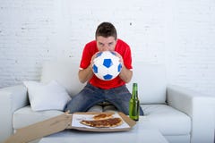 Young man alone holding ball and kissing it in stress wearing team jersey watching football tv Royalty Free Stock Photography Young man alone holding ball and kissing it in stress wearing team jersey watching football tv Royalty Free Stock Photography