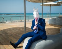 Young man is dressed up a suit under stylish umbrellas on the city waterfront. Unusual traveler in comical mask. Funny unicorn sits at city promenade on Stock Photography Young man is dressed up a suit under stylish umbrellas on the city waterfront. Unusual traveler in comical mask. Funny unicorn sits at city promenade on Stock Photography