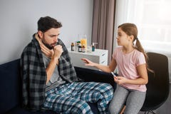 Young man sitting on sofa and coughing. His throat covered with scarf. Blanket is on shoulders. Small girl worry about stock photography Young man sitting on sofa and coughing. His throat covered with scarf. Blanket is on shoulders. Small girl worry about stock photography