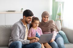 Young muslim parents and girl using laptop together, surfing internet or having video chat with someone, sittig on sofa