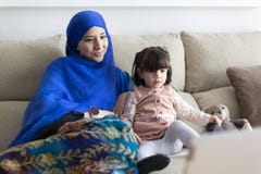 Young Muslim woman with her young daughter at home. Single parent family watching cartoons
