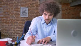 Young nerdy businessman writing notes from laptop, sitting in modern office, serious and concentrated stock video footage Young nerdy businessman writing notes from laptop, sitting in modern office, serious and concentrated stock video footage
