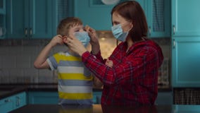 Young 30s mother in medical mask helps kid to wear medical mask on children face at home. Woman and boy in masks looking