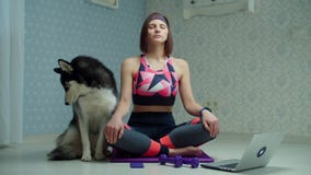 Young 30s woman in sportswear making yoga and meditation exercises on fitness mat with Siberian Husky dog nearby at home