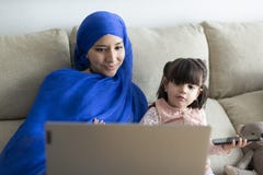 Young single Muslim mother with her young daughter using a laptop at home. Making video call, online entertainment, watching