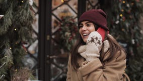 Young smiling attractive woman wearing winter hat and mittens, standing outside and talking on the phone laughing stock video footage Young smiling attractive woman wearing winter hat and mittens, standing outside and talking on the phone laughing stock video footage