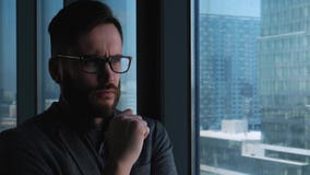 Young successful businessman with a beard thinking in the background of a window overlooking the downtown. High business stock video Young successful businessman with a beard thinking in the background of a window overlooking the downtown. High business stock video