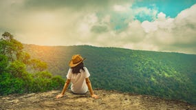 Young traveling woman wearing hat and sitting on the top of the mountain cliff with relaxing mood. Asian woman travel alone.