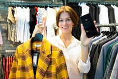 Young Woman Is Holding Black Cellphone And Checkered Coat Of Yellow Color In Clothing Shop For Women Royalty Free Stock Photography Young Woman Is Holding Black Cellphone And Checkered Coat Of Yellow Color In Clothing Shop For Women Royalty Free Stock Photography