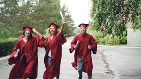 Z podnieceniem kończy studia ucznie jest ubranym togi biegają z dyplomami na kampusu terytorium i tradycyjni kapelusze, ono jest zdjęcie wideo Z podnieceniem kończy studia ucznie jest ubranym togi biegają z dyplomami na kampusu terytorium i tradycyjni kapelusze, ono jest zdjęcie wideo