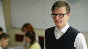Zamyka w górę przystojnego młodego pracownika jest ubranym eyeglasses i flanelową koszula w małym biurze z innym personelem w tle zbiory Zamyka w górę przystojnego młodego pracownika jest ubranym eyeglasses i flanelową koszula w małym biurze z innym personelem w tle zbiory