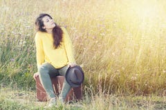 Zmęczona dziewczyna z kapeluszowym obsiadaniem i odpoczywać na brown rocznik walizce w śródpolnej drodze podczas lato zmierzchu S Fotografia Stock Zmęczona dziewczyna z kapeluszowym obsiadaniem i odpoczywać na brown rocznik walizce w śródpolnej drodze podczas lato zmierzchu S Fotografia Stock