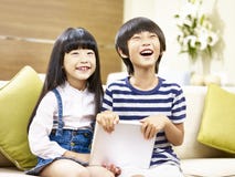 Zwei asiatische Kinder, die auf dem Couchlachen sitzen lizenzfreie stockbilder Zwei asiatische Kinder, die auf dem Couchlachen sitzen lizenzfreie stockbilder
