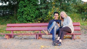 Zwei junge Studenten, die auf einer Parkbank plaudern Genießen Sie Tablette Koreanischer Mann und kaukasische Frau stock video footage Zwei junge Studenten, die auf einer Parkbank plaudern Genießen Sie Tablette Koreanischer Mann und kaukasische Frau stock video footage