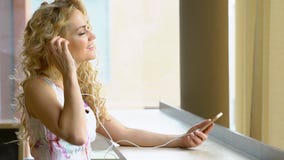 Zwolnione tempo młoda blondynki dziewczyna w sukni jest słuchającym muzyką jej smartphone przy sklep z kawą kawiarnią zbiory Zwolnione tempo młoda blondynki dziewczyna w sukni jest słuchającym muzyką jej smartphone przy sklep z kawą kawiarnią zbiory