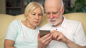 Ältere Paare, die zu Hause auf Sofa sitzen Moderne Pensionäranwendung beweglich, grasend und lesen Nachrichten stock video Ältere Paare, die zu Hause auf Sofa sitzen Moderne Pensionäranwendung beweglich, grasend und lesen Nachrichten stock video