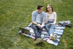 Étudiants souriant tout en lisant et à l'aide de l'ordinateur portable sur l'herbe verte en parc Éducation heureuse photos libres de droits Étudiants souriant tout en lisant et à l'aide de l'ordinateur portable sur l'herbe verte en parc Éducation heureuse photos libres de droits