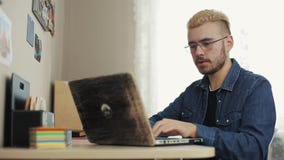 Η άποψη Siade του chherful νέου αρσενικού σπιτιού freelancer με τα γυαλιά και την κίτρινη τρίχα ανοίγει και αρχίζει το lap-top σπ απόθεμα βίντεο Η άποψη Siade του chherful νέου αρσενικού σπιτιού freelancer με τα γυαλιά και την κίτρινη τρίχα ανοίγει και αρχίζει το lap-top σπ απόθεμα βίντεο