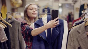 Η αρκετά νέα ευτυχής γυναίκα παίρνει selfie στη κάμερα smartphone με ένα σακάκι από ένα ράφι σε ένα κατάστημα ιματισμού απόθεμα βίντεο Η αρκετά νέα ευτυχής γυναίκα παίρνει selfie στη κάμερα smartphone με ένα σακάκι από ένα ράφι σε ένα κατάστημα ιματισμού απόθεμα βίντεο