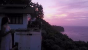 Νέα γυναίκα που στέκεται στο μπαλκόνι, που προσέχει το ηλιοβασίλεμα και που πίνει τον καυτό καφέ με το όμορφο τοπίο βουνών επάνω φιλμ μικρού μήκους Νέα γυναίκα που στέκεται στο μπαλκόνι, που προσέχει το ηλιοβασίλεμα και που πίνει τον καυτό καφέ με το όμορφο τοπίο βουνών επάνω φιλμ μικρού μήκους