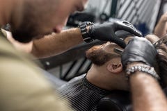 Ο κουρέας στα μαύρα γάντια τακτοποιεί τη γενειάδα του βάναυσου νεαρού άνδρα με ένα ευθύ ξυράφι σε ένα barbershop στοκ εικόνες Ο κουρέας στα μαύρα γάντια τακτοποιεί τη γενειάδα του βάναυσου νεαρού άνδρα με ένα ευθύ ξυράφι σε ένα barbershop στοκ εικόνες