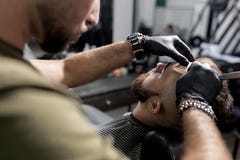 Ο κουρέας στα μαύρα γάντια τακτοποιεί το ναό του βάναυσου γενειοφόρου νεαρού άνδρα με ένα ευθύ ξυράφι σε ένα barbershop στοκ φωτογραφίες με δικαίωμα ελεύθερης χρήσης Ο κουρέας στα μαύρα γάντια τακτοποιεί το ναό του βάναυσου γενειοφόρου νεαρού άνδρα με ένα ευθύ ξυράφι σε ένα barbershop στοκ φωτογραφίες με δικαίωμα ελεύθερης χρήσης