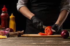 Ο μάγειρας κόβει μια φρέσκια ντομάτα για να κάνει burger, ένα χάμπουργκερ Σε ένα υπόβαθρο με τα συστατικά Εύγευστος και γρήγορο φ στοκ φωτογραφία με δικαίωμα ελεύθερης χρήσης Ο μάγειρας κόβει μια φρέσκια ντομάτα για να κάνει burger, ένα χάμπουργκερ Σε ένα υπόβαθρο με τα συστατικά Εύγευστος και γρήγορο φ στοκ φωτογραφία με δικαίωμα ελεύθερης χρήσης
